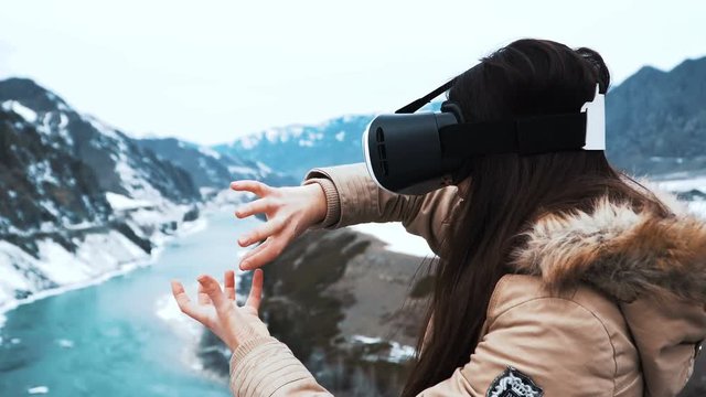 Young girl or teenager uses virtual reality glasses. Solves a puzzle or plays a game. Perhaps creating a design. Enjoys adventure and travel. On a background of winter mountains and a green river