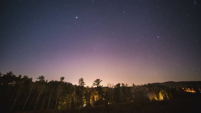 Time lapse of stars above lit forest with shadow moving across trees