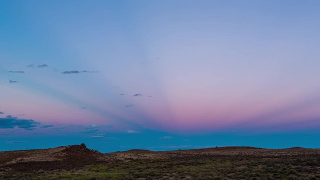 A static timelapse shot at twilight of a typical rocky Karoo landscape against a colorful magenta and blue sky with scattered clouds and light streaks across the sky, as the night falls