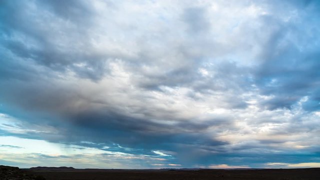 An early morning timelapse before sunrise with blue moody clouds rolling through while tilting down to a vast open rocky landscape with a dirt road going through