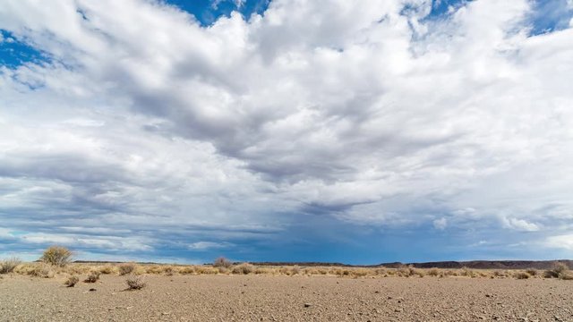 A static timelapse, very low angle, interesting point of viewc of a landscape and lots of foreground with thick clouds against a bright blue sky, gathering in the distance where rain is falling from dark blue clouds