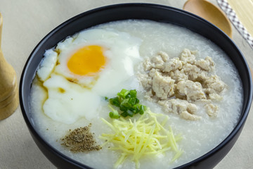 Rice porridge is topping with ginger, spring onion, pepper, chicken, and onsen egg, Breakfast set on wooden table, spoon, pepper grinder. breakfast in Asia call 