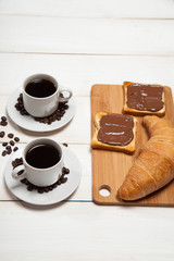 Cups of coffee with a croissant and toast with chocolate on a white wooden table