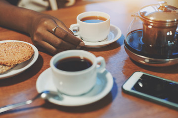 Hand of young black girl touching cup with delicious tea on table with sesame biscuits, teapot, one more cup and smartphone, biracial teenager arm holding white saucer while meeting during breakfast
