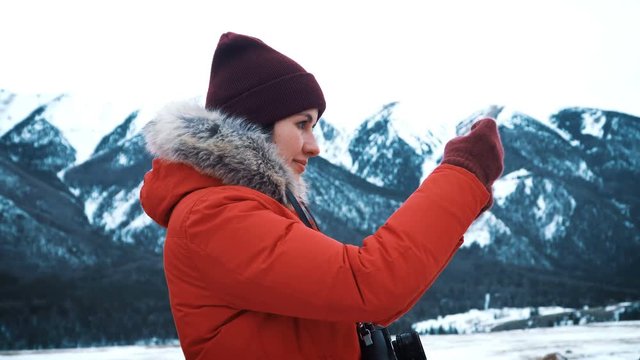Young girl or teen makes selfie or self-portrait on a smartphone against the background of big mountains and the green mountain river. Photographs the winter nature. Enjoys adventure and travel