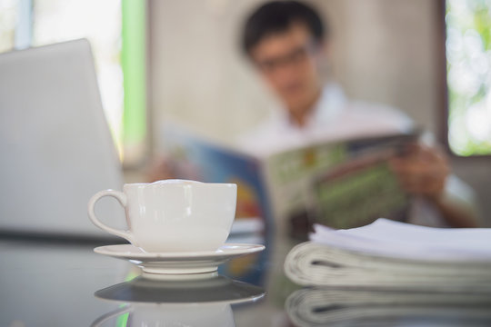 A white cup of coffee on the foreground with elegant young businessman reading newspaper at office blur background.