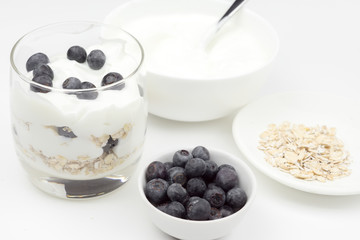 A bowl of blueberries next to a bowl of oatmeal, a bowl of yogurt and glass of the finished dish.