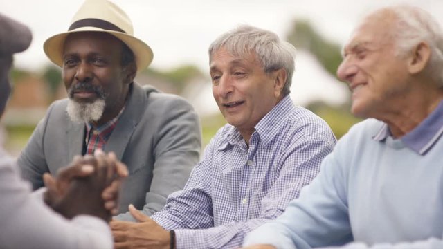  Happy senior male friends chatting & laughing together outdoors in the park