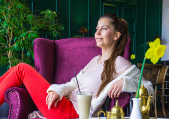 Beautiful smiling girl with long dark hair sitting on a lilac armchair at the nice interior cafe. Fashionably dressed young brunette woman in a modern city bar having a break. 