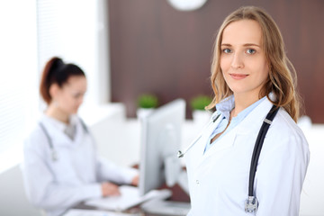 Beautiful young smiling female doctor standing in a hospital with her colleague in the background