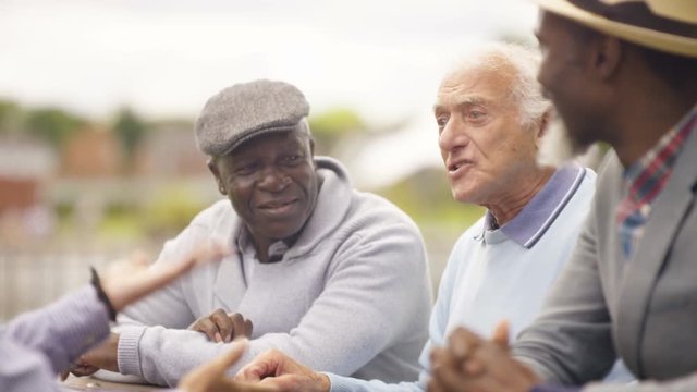  Happy senior male friends chatting & laughing together outdoors in the park