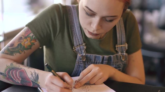 Young attractive red haired woman tattoo artist sitting at table and creating sketch for tattooing in studio indoors