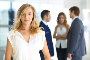 Blonde woman with touchpad computer looking at camera and smiling while business people are shaking hands over office background