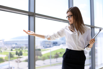 Modern business woman pointing into wide glass window in office while standing and keeping a clipboard. Concept of presentation and meeting.