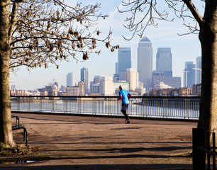 A male jogger in bright sunshine on the Thames Path with Canary Wharf buildings in the distance
