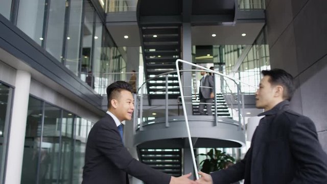  Businessmen meet & shake hands in lobby area of large modern office building