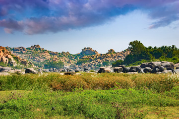 Beautiful landscape with green meadow, unique rock formations of larger rocks and cloudy sky in Hampi, India