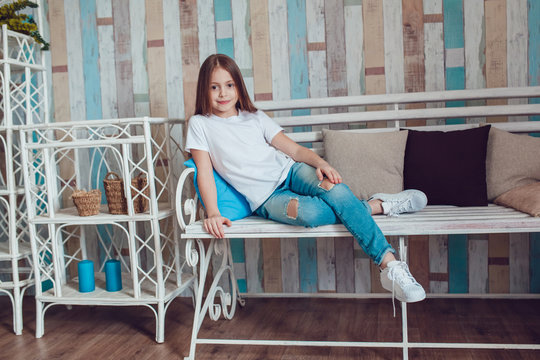 Beautiful little girl in blank white T-shirt and jeans is sitting on a wooden couch.