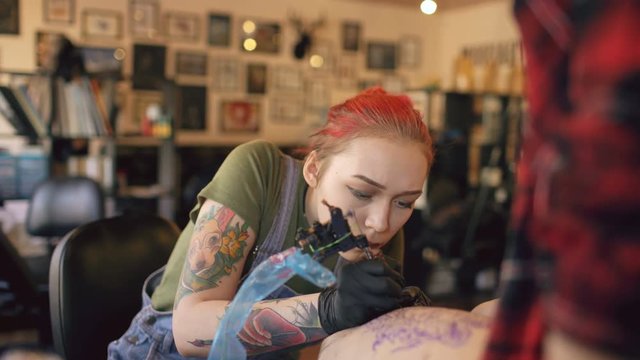 Young female red haired tattoo artist tattooing picture on leg of client over sketch in studio indoors