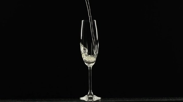 Sparkling yellow water is pouring into stem glass standing on black background in slowmotion