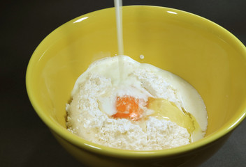 Pouring milk into yellow ceramic mixing bowl with dry flour and egg for making pancakes 
