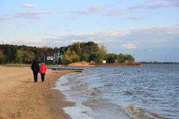 Para ludzi spaceruje z psem po plaży wzdłuż  jeziora.