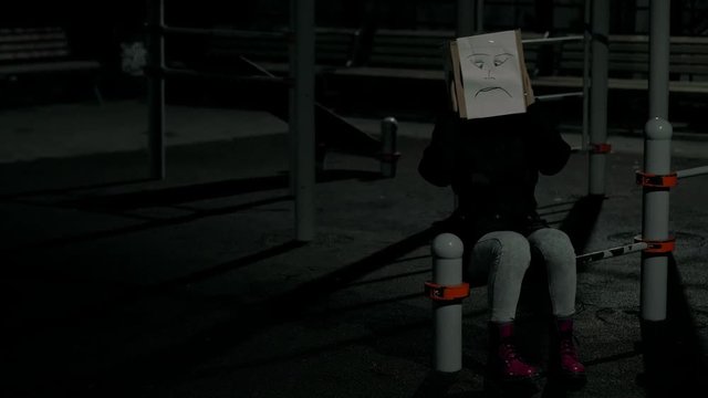 Girl at night wearing a box with an unhappy face drawing on her head. Concept of sorrow and unhappiness. 4K UHD 60 to 24fps