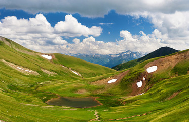 A small lake in the area of alpine meadows in the Caucasus. White clouds in the blue sky.