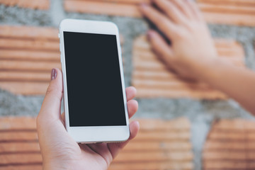 Mockup image of hand holding white mobile phone with blank black screen and brick wall background