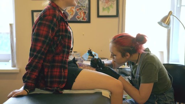 Red haired girl tattoo artist tattooing picture on lef of young girl client in studio