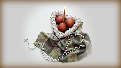 Naklejka premium Easter composition on the table. Colored eggs in a white wicker heart with a burning candle and a cross.