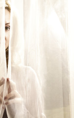 Portrait of blond and attractive woman standing next to the window behind the curtains. Silhouette of beautiful woman with brown eyes.