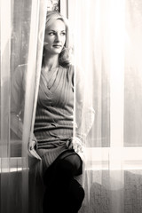 Portrait of blond and attractive woman standing next to the window behind the curtains. Silhouette of beautiful woman with brown eyes.