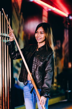 Young beautiful girl wearing leather jacket in a billiard club, with cue stick preparing for the game