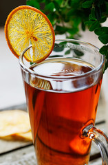 High glass cup of black tea with a slice of dried orange on a bright wooden rustic background  and foliage.