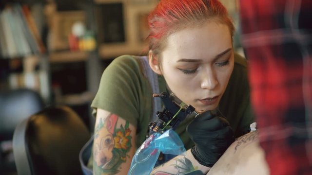 Closeup of beautiful young red haired woman tattoo artist tattooing picture on leg of her girl client in studio indoors