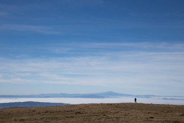A small human figure on top of a mountain, beneath a huge sky, looking at a sea of fog