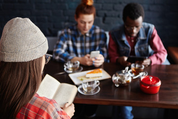 Redhead woman making online order while shopping via internet on mobile telephone while having lunch in modern cafe interior with friends. Selective focus on unrecognizable girl who is reading book