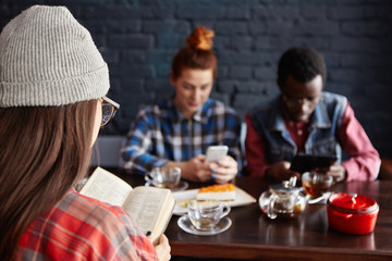 Multiethnic group of stylish young students drinking tea at cafe during break: girl in hat reading book while redhead woman and African man using electronic gadgets. People and modern technologies