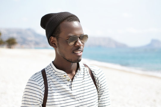 Handsome black hipster wearing stylish hat, sailor shirt, shades and backpack walking alone on urban beach, admiring Maritime seaside landscape while traveling abroad during summer vacations