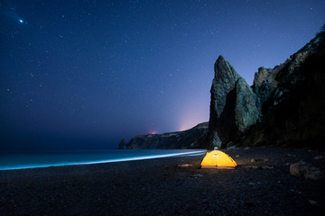 Glowing camping tent on a beautiful sea shore with rocks at night under a starry sky
