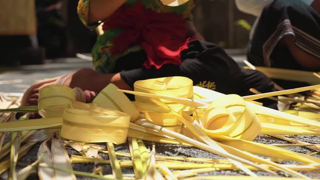 Close up of palm leaf decorations for penjor on Galungan Day (Bali holiday ceremony) made by balinese man and woman sitting behind. Shot with Sony a7s on tripod on sunny day
