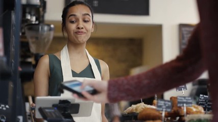  Cheerful worker serving a customer who uses smartphone to pay in coffee shop. - Powered by Adobe