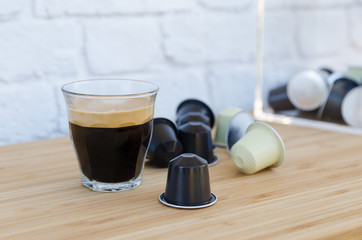 Cup of black coffee with foam and coffee capsules on wooden background.