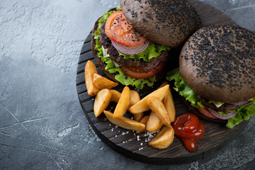 Two black beef burgers with potato wedges on a wooden serving board, horizontal shot, close-up