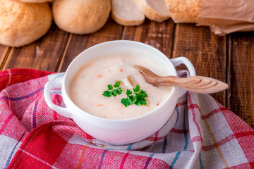 Cream soup in white bowl with wood spoon near bread on wooden background. Rustic style