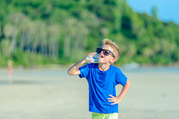 Portrait of a thirsty handsome boy in sunglasses drinking water on the desert tropical beach