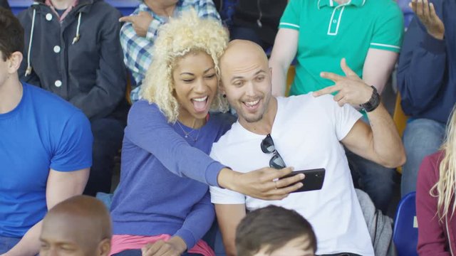  Couple sitting in the crowd at sports event pose to take a selfie with phone