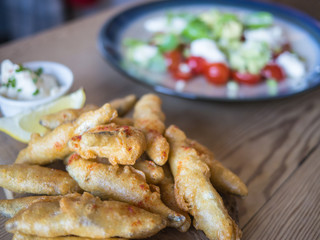 Deep fried whitebait, sprats in batter served with a lemon slice and dip on a wooden table with a tomato, feta cheese and avocado salad.