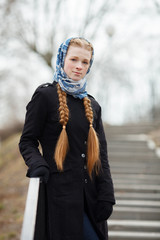 Young beautiful fashionable redhead woman with braids hairdo in blue white headcraft stylish denim black trench jacket posing on stairway in city park spring time